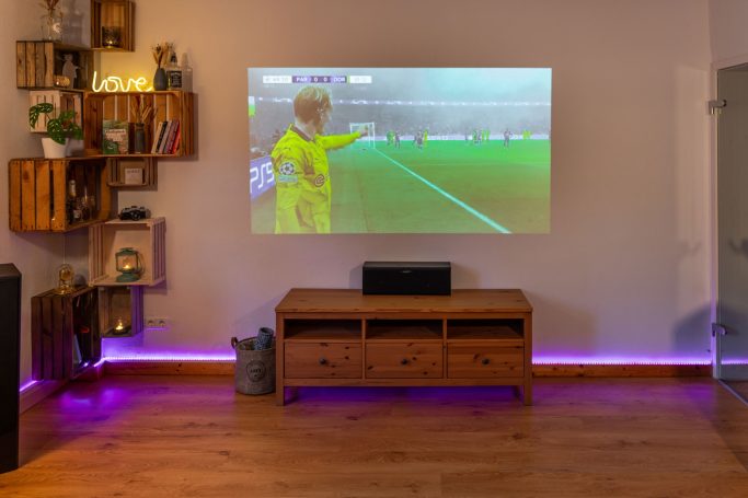 Ein Wohnzimmer mit einem Projektor, der ein Fußballspiel an die Wand projiziert.