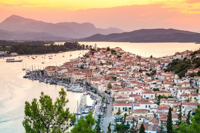 Landschaft mit griechischem Küstendorf, Sonnenuntergang über dem Wasser und Hügeln im Hintergrund.