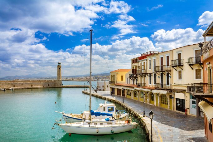 Hafen von Rethymon auf Kreta mit Booten, bunten Gebäuden und Leuchtturm unter bewölktem Himmel.