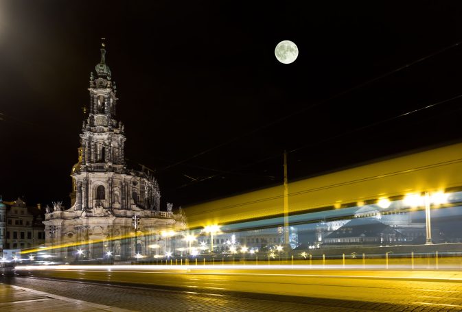Nachtaufnahme der Dresdner Hofkirche mit Vollmond und Lichtspuren von Fahrzeugen.