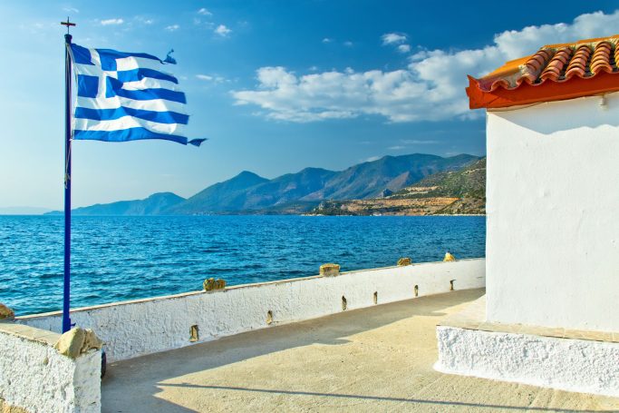 Griechische Flagge weht vor einer Küstenlandschaft mit blauem Wasser und Bergen.