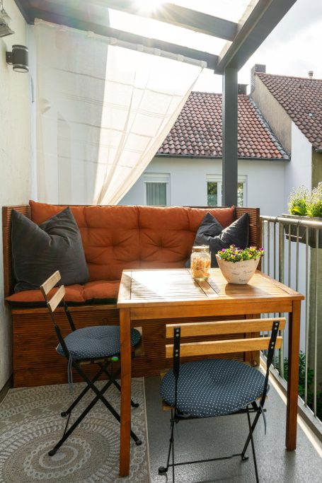 Gemütlicher Balkon mit Holzbank, Tisch und Pflanzen, von Sonnenschein durchflutet.