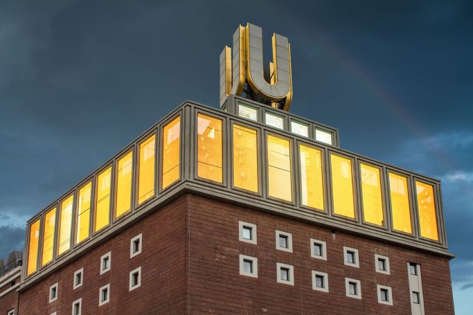 Gebäude mit leuchtenden Fenstern und einem großen "U" oben, vor einem bewölkten Himmel.
