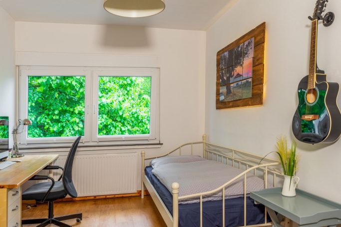 Helles Zimmer mit Bett, Schreibtisch, Stuhl, einem Fenster und einer Gitarre an der Wand.
