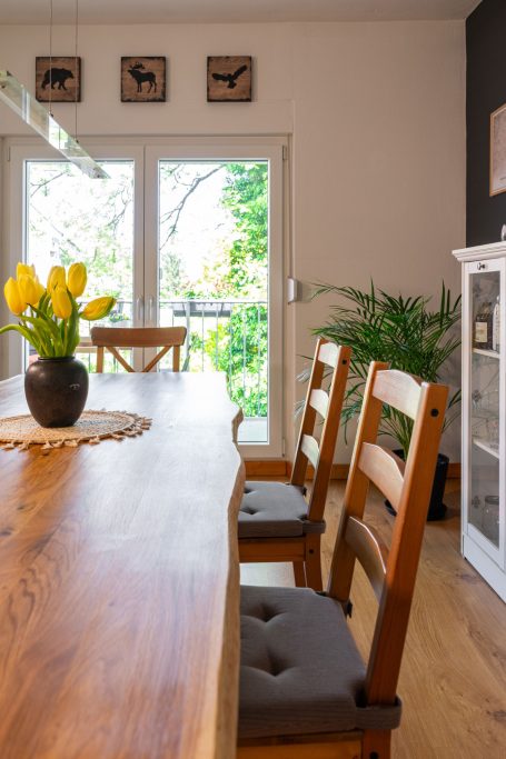 Holztisch mit Blumen und Stühlen, Blick auf eine grüne Terrasse durch Fenster.