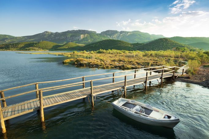 Ein Steg führt zu einem ruhigen See, umgeben von Bergen und Bäumen. Ein Boot liegt am Ufer.